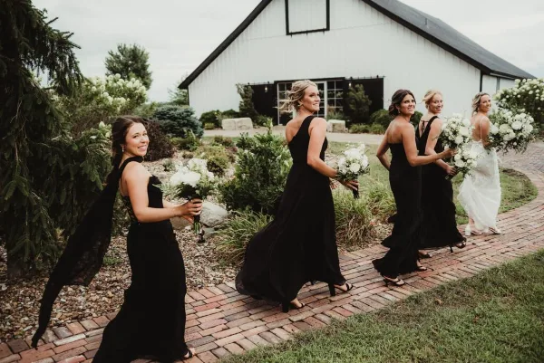 A bride and bridesmaids in black dresses walking on a brick path in front of a white barn.