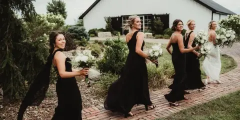 A bride and bridesmaids in black dresses walking on a brick path in front of a white barn.