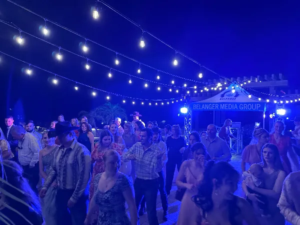 Outdoor wedding reception at night with guests dancing under string lights near a DJ booth labeled Belanger Media Group.