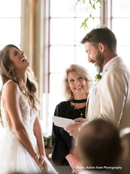 A female wedding officiant in a black top stands between a bride and groom, holding a script and smiling during a ceremony.