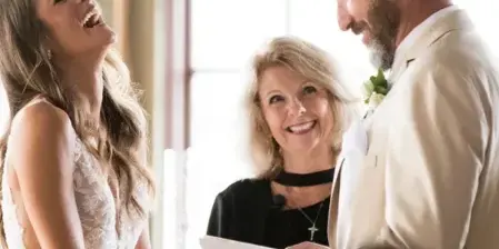 A female wedding officiant in a black top stands between a bride and groom, holding a script and smiling during a ceremony.