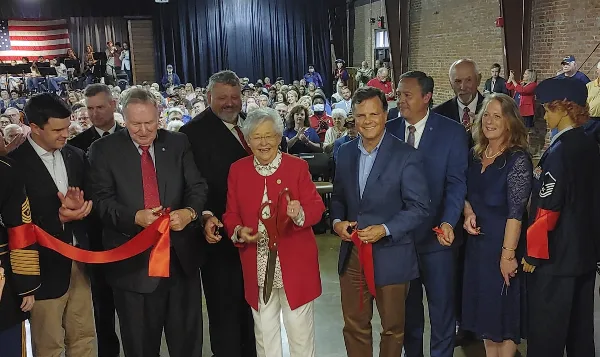 Officials and military personnel cutting a red ribbon during a ceremony in a large event hall with a seated audience.