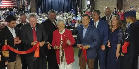 Officials and military personnel cutting a red ribbon during a ceremony in a large event hall with a seated audience.