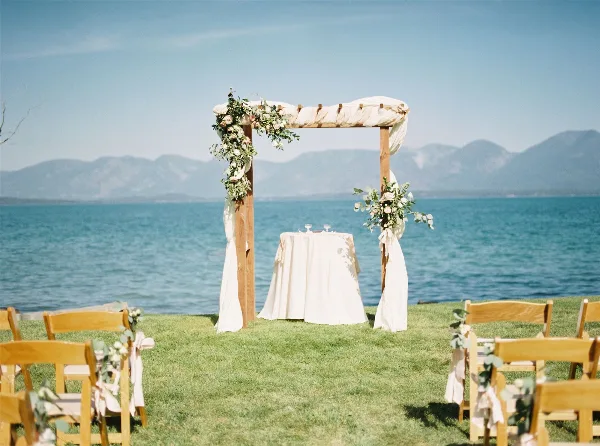 Outdoor wedding ceremony setup with a floral arch, white-draped table, and wooden chairs on a grassy lawn overlooking a lake and mountains.
