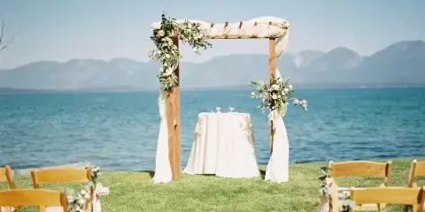 Outdoor wedding ceremony setup with a floral arch, white-draped table, and wooden chairs on a grassy lawn overlooking a lake and mountains.