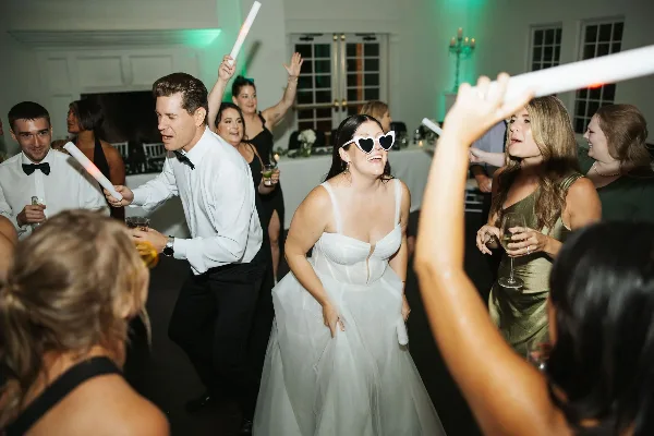 A bride and wedding guests dancing at a reception with glow sticks and green uplighting.