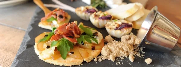 A close-up photograph of a gourmet appetizer featuring a tartine topped with prosciutto and fresh herbs, served on a dark slate board.