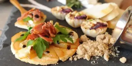 A close-up photograph of a gourmet appetizer featuring a tartine topped with prosciutto and fresh herbs, served on a dark slate board.
