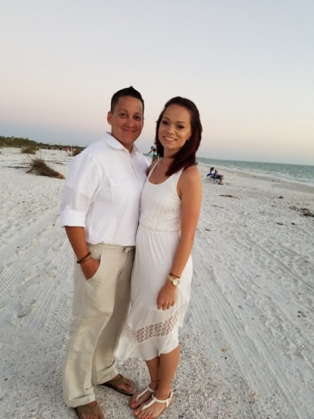 A couple in white wedding attire standing on a sandy beach.