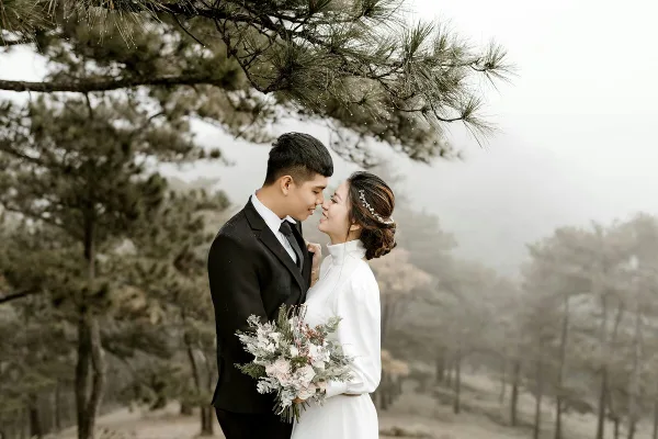 A bride and groom standing together in a misty, wooded outdoor setting, with the bride holding a bouquet.