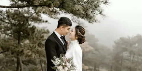 A bride and groom standing together in a misty, wooded outdoor setting, with the bride holding a bouquet.
