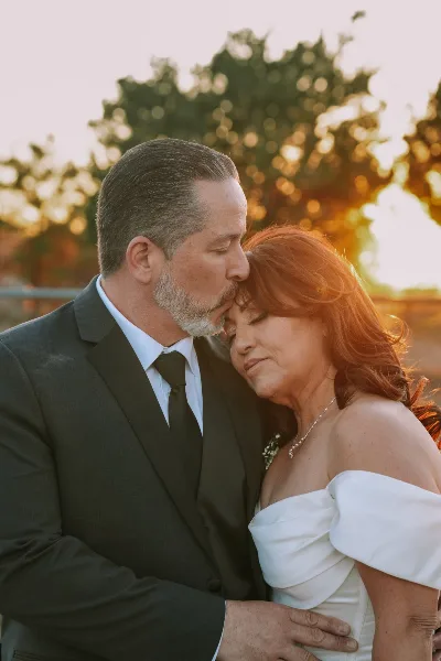 A groom in a dark suit kisses the forehead of a bride in a white off-the-shoulder wedding dress during a sunset outdoor wedding portrait.