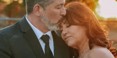 A groom in a dark suit kisses the forehead of a bride in a white off-the-shoulder wedding dress during a sunset outdoor wedding portrait.