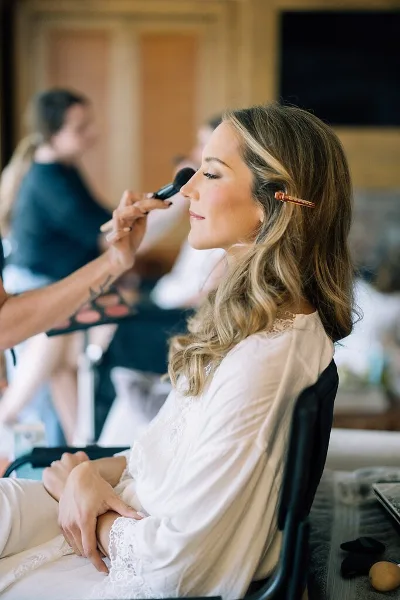 A makeup artist applying makeup to a bride in a white lace robe during a wedding preparation session.