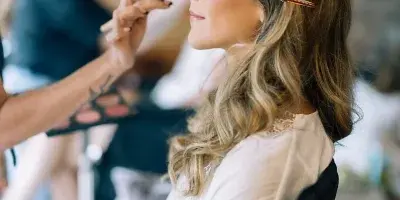 A makeup artist applying makeup to a bride in a white lace robe during a wedding preparation session.