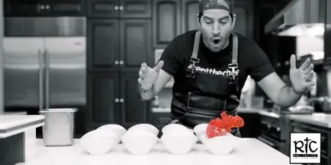 Chef Jason Febres in a professional kitchen wearing a Rent The Chef apron, standing behind a counter with white bowls and a red garnish.