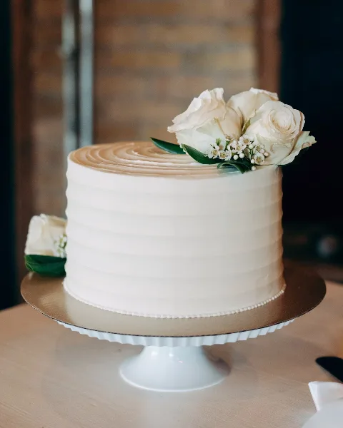A white frosted cake decorated with fresh white roses and baby's breath on a gold cake stand.