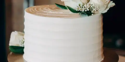A white frosted cake decorated with fresh white roses and baby's breath on a gold cake stand.