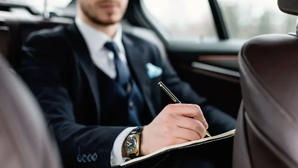 Professional chauffeur in a suit and tie sitting in the back seat of a luxury car, writing in a notebook.