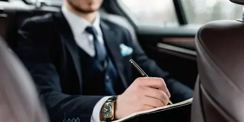 Professional chauffeur in a suit and tie sitting in the back seat of a luxury car, writing in a notebook.