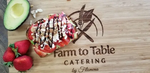 A wooden cutting board featuring a slice of bruschetta topped with strawberries, avocado, and nuts, next to fresh strawberries and an avocado half, with the Farm to Table Catering logo.