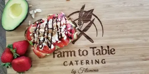 A wooden cutting board featuring a slice of bruschetta topped with strawberries, avocado, and nuts, next to fresh strawberries and an avocado half, with the Farm to Table Catering logo.
