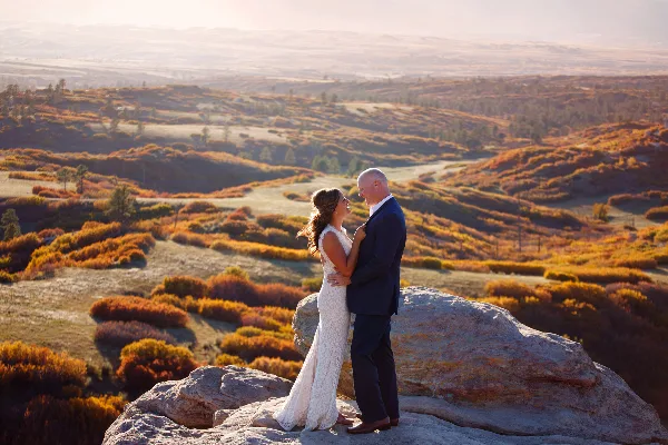 A bride and groom standing on a rocky outcrop overlooking a vast, rolling landscape with autumn foliage.