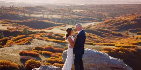 A bride and groom standing on a rocky outcrop overlooking a vast, rolling landscape with autumn foliage.