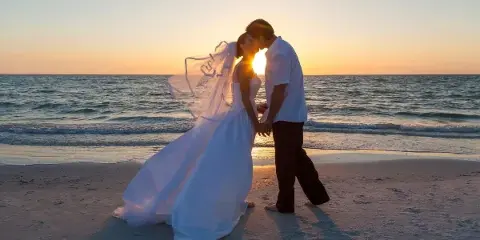 A bride and groom standing on a sandy beach at sunset with the ocean in the background.