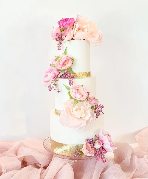 A three-tiered white wedding cake decorated with pink peonies and gold bands, sitting on a glass stand.