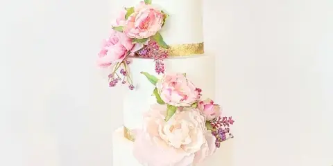 A three-tiered white wedding cake decorated with pink peonies and gold bands, sitting on a glass stand.