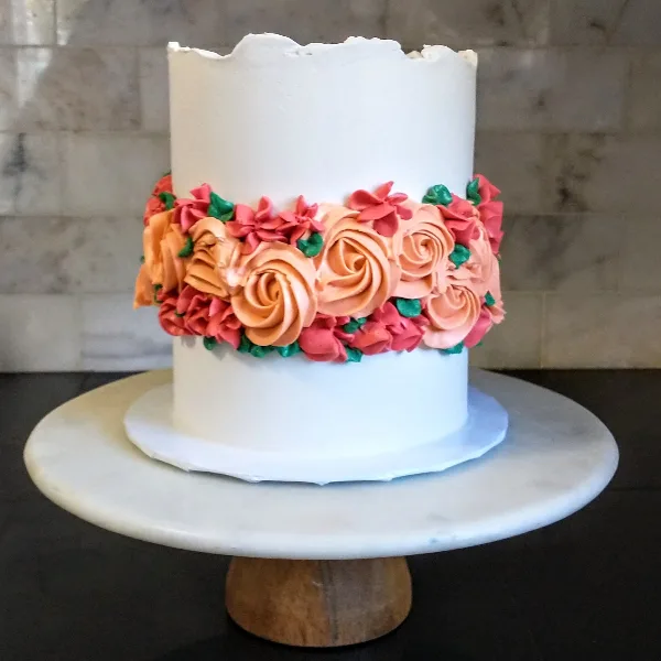 A white frosted cake decorated with a band of orange and pink buttercream rosettes and green leaves, sitting on a white marble cake stand.