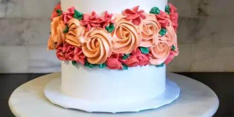 A white frosted cake decorated with a band of orange and pink buttercream rosettes and green leaves, sitting on a white marble cake stand.