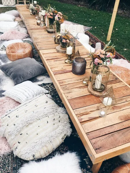 Outdoor event setup featuring a long wooden table decorated with candles, flowers, and lanterns, surrounded by floor cushions and pillows on a rug.