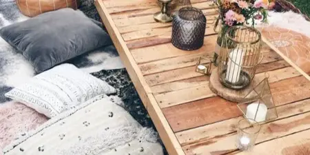 Outdoor event setup featuring a long wooden table decorated with candles, flowers, and lanterns, surrounded by floor cushions and pillows on a rug.