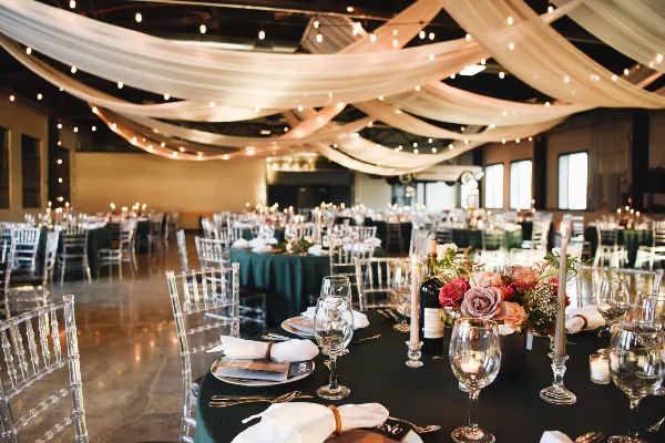 A spacious event venue interior featuring white ceiling draping, string lights, and round tables set with dark green linens, clear chairs, and floral arrangements.
