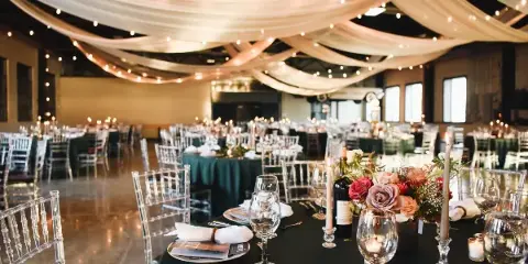 A spacious event venue interior featuring white ceiling draping, string lights, and round tables set with dark green linens, clear chairs, and floral arrangements.