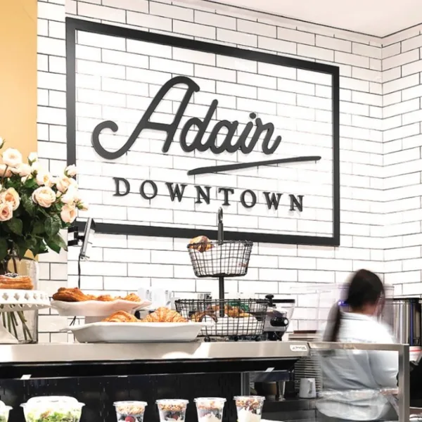 Interior of Adair Downtown café with a white subway-tile wall, framed 'Adair Downtown' sign, pastries on the counter, and a vase of flowers.