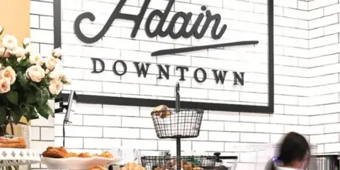 Interior of Adair Downtown café with a white subway-tile wall, framed 'Adair Downtown' sign, pastries on the counter, and a vase of flowers.