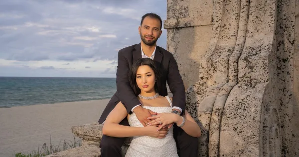 A professional wedding photograph of a couple posing together on a beach with a stone structure in the background.