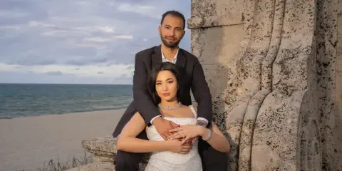 A professional wedding photograph of a couple posing together on a beach with a stone structure in the background.