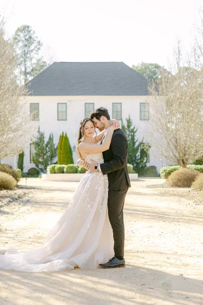 A bride and groom embracing in a romantic portrait in front of a white estate house.