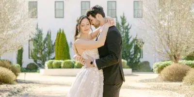 A bride and groom embracing in a romantic portrait in front of a white estate house.