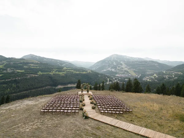 A wedding ceremony setup featuring a floral arch and rows of chairs on a grassy hillside overlooking a mountain valley.