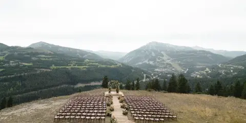 A wedding ceremony setup featuring a floral arch and rows of chairs on a grassy hillside overlooking a mountain valley.