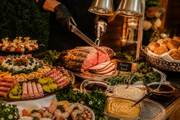 Catering table with roast beef being carved, charcuterie, salads, rolls, and sauces under heat lamps