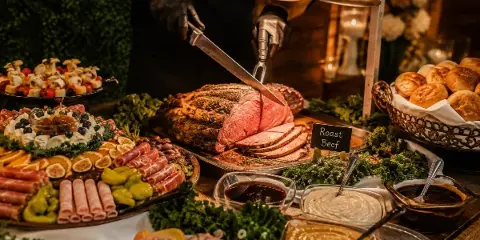 Catering table with roast beef being carved, charcuterie, salads, rolls, and sauces under heat lamps