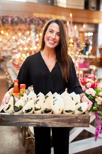 A catering staff member holding a wooden tray of wrapped food items at a wedding reception.