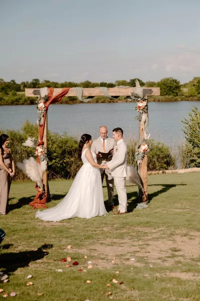 Bride and groom exchanging vows under a rustic floral arch on a lawn overlooking a lake.