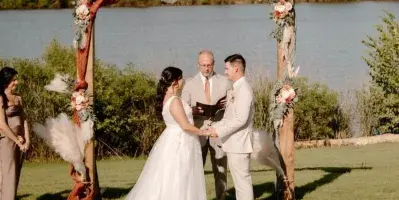 Bride and groom exchanging vows under a rustic floral arch on a lawn overlooking a lake.